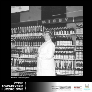 Kobieta stojąca bokiem w sklepie ubrana w biały fartuch, uśmiecha się. W tle stoją regały z napisem miody, na pólkach poustawiane są butelki z winami. Zdjęcie czarno-białe wykonane w latach 50/60 tych.