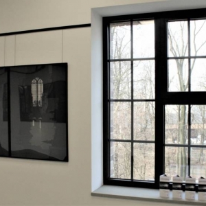 Na ścianie zawieszone są trzy czarno-białe fotografie w czarnych ramach. Ze zdjęć wyłaniają się witraże z Katedry św. Stanisława i św. Wacława. Obok ściany widoczne jest okno Galerii na Piętrze, za którym widać drzewa z pobliskiego parku.