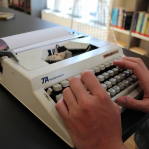 Na stole stoi stara maszyna do pisania, z włożoną czystą kartką papieru. Na klawiszach maszyny położone są męskie dłonie.