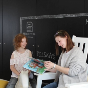 Roześmiana kobieta w kręconych, rozwianych włosach czyta książkę dla swojej córki, która również szeroko się uśmiecha. Dziewczynka w wieku 10 lat siedzi na bujanym koniku. W tle są czarne szafki na których kredą namalowana jest telewizor, w którym znajduje się napis Cała Polska Czyta dzieciom, MiniTv oraz logo biblioteki w Żorach. 