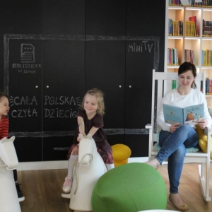 W strefie dla dzieci na bujanym fotelu siedzi uśmiechająca się kobieta, która czyta książkę dzieciom. Obok niej na bujanych konikach siedzą dzieci w wieku 3-4 lat. Dzieci uśmiechają się. W tle jest regał z książkami dla dzieci, czarne szafki na których kredą namalowany jest telewizor, w którym znajduje się napis Cała Polska Czyta dzieciom, MiniTv oraz logo biblioteki w Żorach. 
