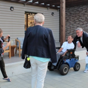 Cztery osoby stoją na świeżym powietrzu. Mężczyzna pozuje do zdjęcia z mężczyzną z niepełnosprawnością ruchową siedzącym na wózku inwalidzkim. 