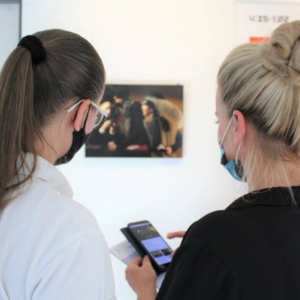 Dwie kobiety odwrócone tyłem, spoglądają w ekran telefonu, w którym widoczne jest rozmazane zdjęcie i tekst. To samo zdjęcie wisi na ścianie przed nimi.
