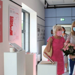 Trzy kobiety w różnym wieku rozmawiają ze sobą. Po lewej stronie widoczne są zawieszone na ścianie grafiki oraz eksponaty umieszczonych na słupkach wystawowych. Jedna z kobiet trzyma bukiet róż.