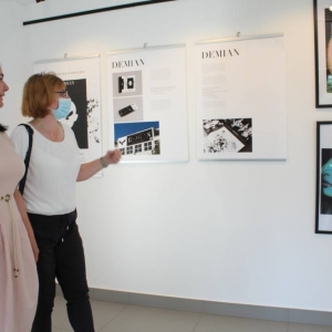 Dwie kobiety idąc przez galerię rozmawiają i spoglądają na prace graficzne zawieszone na ścianie. Na planszach znajduje się opis prac oraz zdjęcia. Z prawej strony, wiszą dwie grafiki przedstawiające kolorowe twarze młodych mężczyzn. 