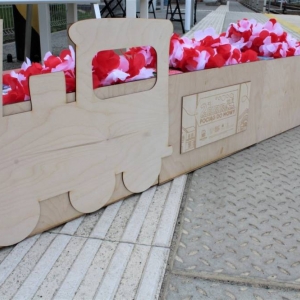 Na chodniku przed torami kolejowymi sto drewniany pociąg. Składa się z dwóch wagonów. Na pierwszym wycięta jest lokomotywa, oraz na jednym z wagonów jest wycięta drewniana tabliczka z logo projektu Pociąg do Mowy 2. W pociągu umieszczone są sztuczne kwiatowe girlandy oraz książki.