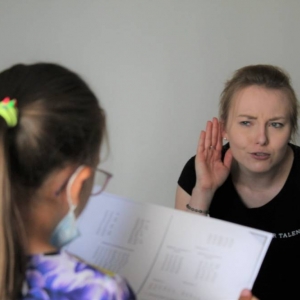 Kobieta instruktorka nachyla się do stojącej przed nią dziewczynki. Przykłada rękę do ucha chcąc usłyszeć, co odczytuje z kartki dziewczynka.