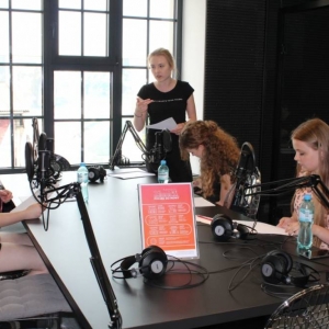 Wnętrze studia radiowego. Przy stole i mikrofonach siedzą trzy dziewczyny, trzymają kartki. Nad nimi, po prawej stronie stoi instruktorka, która gestykuluje i coś im tłumaczy.