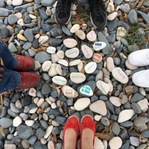 Zdjęcie czterech par nóg i butów stojących na kamieniach, wśród których leżą pomalowane kamienie.