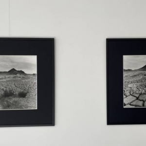 Na ścianie zawieszone są dwie fotografie w kwadratowych ramach. Zdjęcia przedstawiają popękaną ziemię, a w tle góry usypane przez człowieka.