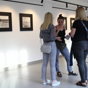 Trzy kobiety po 30-stce rozmawiają w przestrzeni galerii. Wszystkie są ubrane swobodnie rozmawiają, jedna uśmiecha się spoglądając na fotografującego. Na ścianach zawieszone są fotografie w kwadratowych ramach przedstawiające podróż do Azerbejdżanu. 