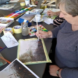 Zdjęcie przedstawia starsze kobiety. Przed nimi na stole położone są farby i kredki akwarelowe, pędzle, taśmy papierowe i naczynie z wodą. Kobiety trzymają pędzle i malują krajobrazy ze zdjęć.
