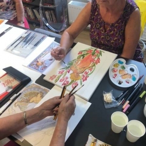 Kobieta w wieku 60 lat w w okularach siedzi przy stoliku i maluje obraz. Na przeciwko niej siedzi przy stole jakaś osoba (widoczne tylko ręce). Wokół nich rozłożone są różne akcesoria plastyczne.