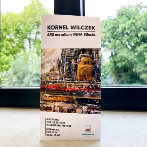 Na parapecie wyeksponowana jest broszurka dotycząca wystawy ARS Metallum VENA Silesia Kornela Wilczka. Przedstawia czas trwania wystawy oraz datę wernisażu. W tle za oknem widać okryte liśćmi drzewa. 