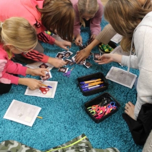 Ujęcie z góry na dywan, na którym kuca kilka dziewczynek wieku 10-13 lat. Pochylają się nad karteczkami oraz wydrukowanymi kartami do gry. Na  gęstym dywanie w pudełkach leżą nożyczki oraz długopisy. 