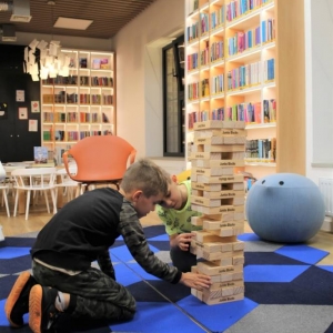 W Strefie Dzieci pomiędzy regałami z książkami dla dzieci, na dywanie klęczy dwóch chłopców w wieku 8-9 lat. Układają wysoką wieżę z klocków jenga.