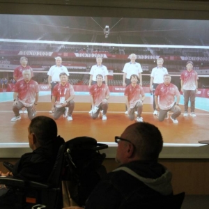 Na ekranie na ścianie wyświetlone jest zdjęcie przedstawiające boisko siatkarskie, na którym stoją zawodnicy oraz międzynarodowi sędziowie, w tym Wojciech Maroszek. Zdjęcie zostało wykonane w Tokio podczas Olimpiady 2020. Przed ekranem widoczni są dwaj mężczyźni, publiczność. Mężczyzna po lewej stronie siedzi na wózku inwalidzkim.