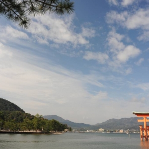 Na wodzie stoi stara, japońska drewniana architektoniczna zabudowa. W tle widoczne są góry porośnięte drzewami oraz budynki. Z przodu zdjęcia, po prawej stronie widoczny jest kawałek iglastego drzewa.