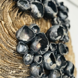 Zbliżenie na tekturę, która tworzy półokręgi z tektury które w ostatecznej formie zamykają się w kole. Na tekturze umieszczone są artystyczne odniesienie do małży i kwiatów pokryte perłowo czarnym szkliwem.