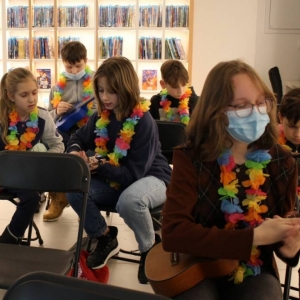 Zdjęcie przedstawia 6 osób, które mają zawieszone na szyi hawajskie kwiaty. Grupa siedzi w pomieszczeniu i w rękach każdy trzyma ukulele. W tle widoczny jest regał z podświetlonymi pólkami na których ułożone są płyty DVD. Osoba na pierwszym planie ma założoną higieniczną maseczkę na twarzy.