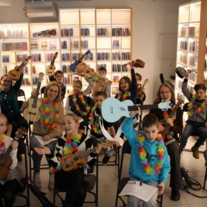 Zdjęcie przedstawia grupę 15 osób siedzących w pomieszczeniu na krzesłach i trzyma ukulele. Jedna kobieta stoi z tyłu z podniesionym nad głową ukulele. Wszyscy na szyi mają zawieszone kwiaty hawajskie. Z tyłu w tle widoczne są oświetlone regały z płytami DVD i audiobookami.