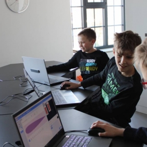 Fragment pomieszczenia. Zbliżenie na trzech chłopców siedzących przy stole mających przed sobą otwarte laptopy. Uśmiechają się. W tle widać okno, fragment zegara. 