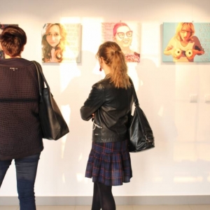 Fragment ściany, na której zawieszone są cztery grafiki przedstawiające kobiety wraz z tekstem. Przed grafikami stoją odwrócone tyłem do fotografa dwie kobiety, które oglądają prace Marty Frej. 