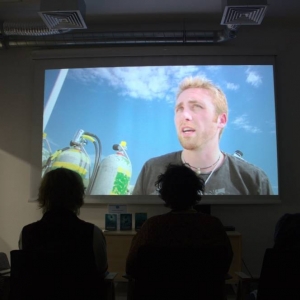 Fragment pomieszczenia na którym wyświetlany jest film. Przed ekranem siedzi czwórka osób i spogląda na duży ekran. Na dużym ekranie uchwycony jest fragment filmu. Mężczyzna mówi, obok niego stoją butle z tlenem.