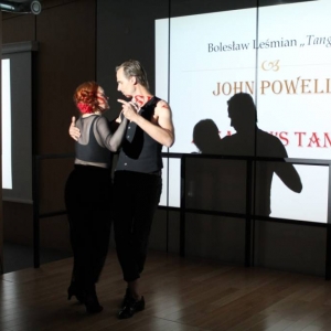 Fragment sali konferencyjnej. W tle wyświetlany jest fragment prezentacji z napisem: Bolesław Leśmian "Tango", John Powell, Assassins's Tango. Na drewnianym podeście na podwyższeniu tańczy dwójka osób argentyńskie tango. Trzymają się w objęciach.  Ich cienie padają na wyświetlającą się za nimi prezentację.