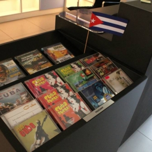 Fragment Strefy Multimediów. Zbliżenie na półkę na której wyeksponowane są płyty CD i DVD z muzyką i filmem kubańskim. Pomiędzy nimi na patyczku stoi flaga Kuby.
