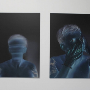 Fragment ściany, na której zawieszone są dwa zdjęcia w negatywie. Fotografie wydrukowane są na płytach PCV i przedstawiają dwóch mężczyzn. Na jednym zdjęciu mężczyzna jest rozmazany i w oddali. Na drugim widać go wyraźnie. Ma oplecione siatką twarz i ręce.