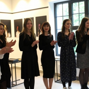 Fragment galerii, na tle ścian z pracami artystycznymi stoi osiem młodych kobiet w wieku 22-23 lat. Są ubrane elegancko w sukienki w stonowanych kolorach. Jedna z kobiet uśmiecha się i mówi do mikrofonu, reszta bije brawa.