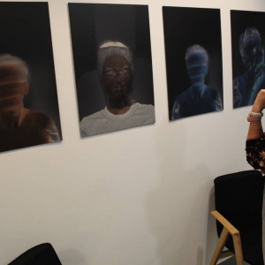 Fragment ściany na której zawieszone są wydrukowane na płytach PCV zdjęcia osób. Są to portrety osób w negatywie  którzy mają pozakrywaną twarz. Pracą przygląda się kobieta w wieku 30 lat z włosami upiętymi w okularach. Rękę trzyma pod brodą.