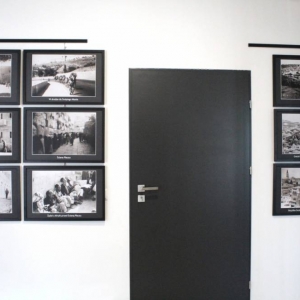 Na zdjęciu widoczne są fotografie w ramach powieszone we wnętrzu. W sumie jest 9 fotografii. Pomiędzy nimi widoczne są drzwi.
