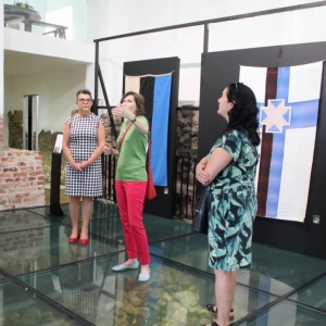 Fragment muzeum. Przy wyeksponowanych flagach Estonii na szklanej podłodze stoją cztery kobiety. Pod podłogą widać fragmenty średniowiecznych murów. Jedna z kobiet opowiada, gestykulując.