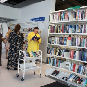 Fragment wnętrza biblioteki. Na regałach i wózku wyeksponowane są książki. Grupa osób spaceruje przy regałach. Jedna z kobiet trzyma notes w ręku i notuje w nim.