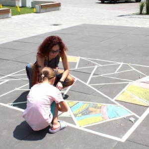 Na płytkach chodnikowych przyklejona jest taśma w romby, kwadraty, trójkąty. W trójkątach malują kolorową kredą dzieci wraz z dziewczyną około 20-letnią.  