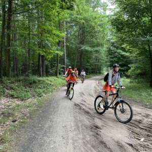 Ujęcie na kilku rowerzystów przejeżdżających przez las. Są wśród nich kobiety, mężczyźni i dzieci. niektórzy mają przyozdobione rowery, niektórzy ubrani są na pomarańczowo. Jadą szutrową drogą, a dookoła widoczne są duże, zielone drzewa.