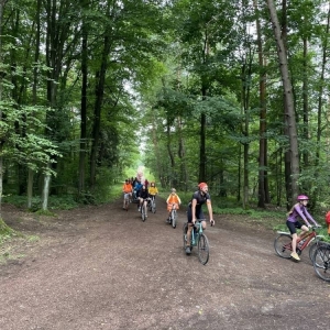Ujęcie na grupę kilkunastu rowerzystów przejeżdżających przez las. Są wśród nich kobiety, mężczyźni i dzieci. niektórzy mają przyozdobione rowery, niektórzy ubrani są na pomarańczowo. Jadą szutrową drogą, a dookoła widoczne są duże, zielone drzewa.
