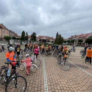 Ujęcie na dużą grupę rowerzystów zlokalizowanych na rynku w Żorach. W ich strojach dominuje kolor pomarańczowy. Wszyscy stoją przy rowerach, część ze sobą rozmawia. W tle widoczna jest karuzela Żorzanka.