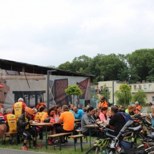 Zbliżenie na Letnią Czytelnię, gdzie na ławeczkach przy stołach siedzą rowerzyści. Większość jest ubrana w pomarańczowe stroje. W tle widoczne są drzewa i zabudowania.