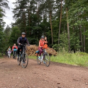 Ujęcie na kilku rowerzystów, którzy szutrową drogą jadą przez las. Machają do fotografa, dookoła widoczne są wysokie, zielone drzewa.