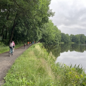 Ujęcie na kilkunastu rowerzystów przejeżdżających przez las. Są oddaleni, widać ich tyły. Jadą szutrową drogą, po lewej stronie widoczne są duże, zielone drzewa, a po prawej fragment jeziora.