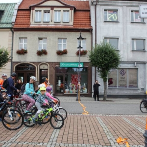 Ujęcie na kilkunastu rowerzystów ruszających ze startu. Są wśród nich kobiety, mężczyźni i dzieci. niektórzy mają przyozdobione rowery, niektórzy ubrani są na pomarańczowo. W tle widoczne są kamienice przy rynku.