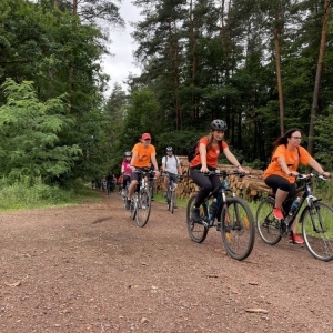 Ujęcie na kilkunastu rowerzystów przejeżdżających przez las. Są wśród nich kobiety, mężczyźni i dzieci. niektórzy mają przyozdobione rowery, niektórzy ubrani są na pomarańczowo. Jadą szutrową drogą, a dookoła widoczne są duże, zielone drzewa.