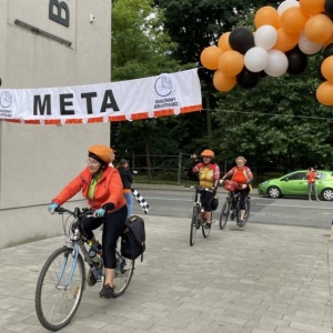 Grupa kilku rowerzystów przejeżdża pod dużym napisem meta. Zawieszony materiał przyozdobiony jest kolorowymi balonami. Część z rowerzystów macha i uśmiecha się. W tle widoczny jest park miejski.