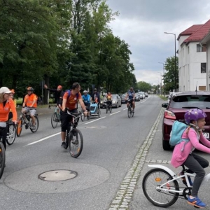 Ujęcie na kilku rowerzystów przejeżdżających główną drogą wśród zabudowań. Są wśród nich kobiety, mężczyźni i dzieci. niektórzy mają przyozdobione rowery, niektórzy ubrani są na pomarańczowo. Uśmiechają się, pokonując ostatni zakręt przed metą.