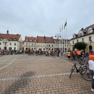 Ujęcie szerokokątne na Rynek w Żorach. Widać zgormadzony tłum rowerzystów. Ustawili się w długiej kolejce do starty. W tle widoczne są stare kamienice przy rynku. Pogoda jest trochę pochmurna.