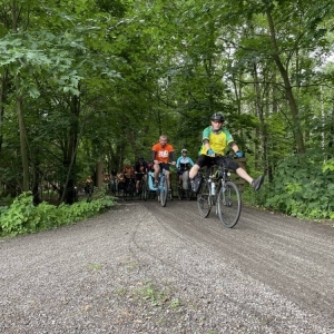 Ujęcie na kilku rowerzystów przejeżdżających przez las. Są wśród nich kobiety, mężczyźni i dzieci. niektórzy mają przyozdobione rowery, niektórzy ubrani są na pomarańczowo. Jadą szutrową drogą, a dookoła widoczne są duże, zielone drzewa. Mężczyzna na pierwszym planie specjalnie do zdjęcia rozszerzył nogi, przyjmując śmieszną pozycję na rowerze.