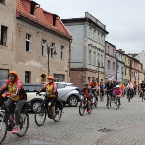 Ujęcie na kilkunastu rowerzystów przejeżdżających przez ulice starego miasta. Są wśród nich kobiety, mężczyźni i dzieci. niektórzy mają przyozdobione rowery, niektórzy ubrani są na pomarańczowo. W tle widoczne są stare kamienice.